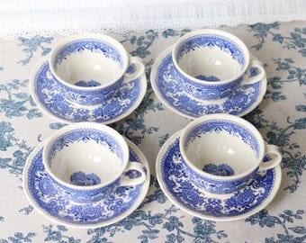 Może przedstawiać: Cztery ceramiczne filiżanki do herbaty z pasującymi spodkami, ozdobione niebiesko-białym wzorem kwiatowym. Każda filiżanka ma zaokrąglone ucho i falisty brzeg, a skomplikowane motywy botaniczne zdobią zewnętrzną część naczyń oraz spodki.