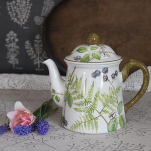 May include: White ceramic teapot with a lid, decorated with green fern and blackberry illustrations. The handle and lid knob have a brown and green finish. A pink and purple flower arrangement is in the foreground.