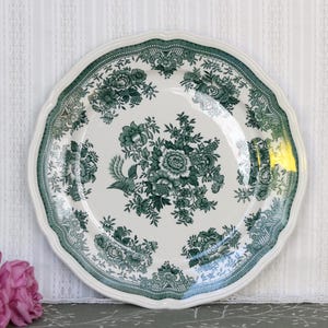 May include: A round ceramic plate with a scalloped edge featuring a dark green floral and bird transferware pattern on a cream background. The design includes a central bouquet of flowers and birds surrounded by a decorative border of foliage.