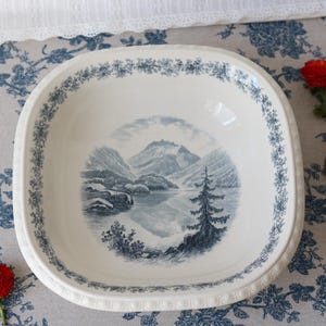 Może przedstawiać: Kwadratowa ceramiczna miska z zaokrąglonymi rogami, posiadająca białą bazę oraz niebieską ilustrację krajobrazu górskiego jeziora i drzewa iglastego w centrum, otoczoną niebieskim wzorem kwiatowym wzdłuż krawędzi naczynia.