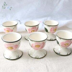 May include: A set of six cream-colored ceramic egg cups, each featuring a hand-painted pink flower with a yellow center and green leaves. The cups have a flared base and a dark green rim, arranged in two rows on a textured off-white surface.