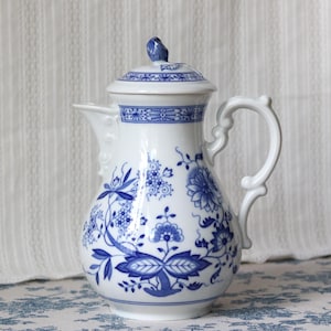 May include: A white porcelain coffee pot featuring a blue floral pattern, a curved handle, and a matching lid with a small knob. The body is decorated with intricate blue botanical designs, including stylised flowers and leaves, set against a smooth, glossy white surface.
