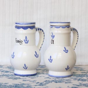 Puede incluir: Dos vinagreras de cerámica blanca con patrones de hojas pintados en azul y bordes azules. Una pieza tiene la palabra Essig y la otra la palabra Öl escritas en negro. Cada recipiente cuenta con un asa, un cuerpo redondeado y una tapa a juego.