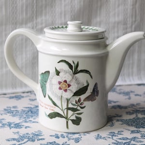 Può includere: Teiera in ceramica bianca con coperchio e manico. La teiera presenta un design floreale e a farfalla con accenti verdi, bianchi e viola. Ha un beccuccio curvo e un piccolo pomello sul coperchio.