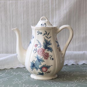 Puede incluir: Tetera de cerámica color crema con tapa, con un diseño floral en rosa, azul y verde. La tetera tiene un pico curvo y un asa grande. El patrón floral incluye rosas y hojas.