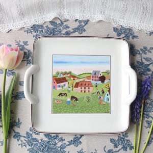 May include: Square white serving tray with a village scene illustration, brown trim, and white handles. Pink and purple flowers are arranged around the tray on a floral tablecloth. The tray's dimensions are not visible.