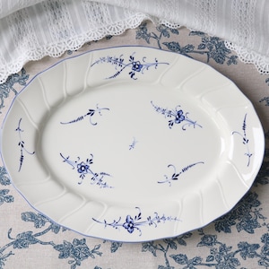 Op de afbeelding: Een ovale, witte keramische serveerschaal met een blauw bloemmotief en een geschulpte rand. De schaal is versierd met blauwe bloemmotieven en een blauwe rand. De schaal staat op een tafelkleed met bloemenpatroon.