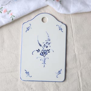 May include: White ceramic chopping board with a blue floral design and a hole for hanging. The board has a scalloped top edge and a blue border. The floral design features a central flower and delicate leaves.
