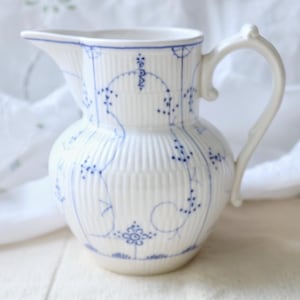 Könnte beinhalten: Weißer Keramikkrug mit blauem Blumen- und Rankendesign. Der Krug hat einen Henkel und einen Ausguss. Der Körper des Kruges ist vertikal gerippt. Das blaue Design umfasst kleine Punkte und Blumenmuster. Der Krug dient wahrscheinlich zum Servieren von Getränken.