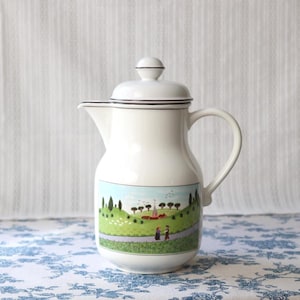 Op de afbeelding: Witte keramische koffiepot met deksel en handvat. De pot heeft een landschap met een weg, bomen en figuren. De rand en het deksel zijn geaccentueerd met een dunne zwarte lijn. De pot staat op een blauw-wit patroon oppervlak.