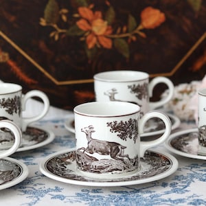 Può includere: Un set di tazzine da caffè in ceramica bianca con piattini abbinati, caratterizzato da una stampa monocromatica marrone che raffigura un cervo che salta in un paesaggio boschivo. Ogni tazzina ha un manico curvo e un bordo liscio.