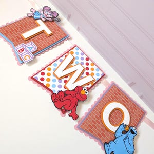 May include: A decorative banner spelling the word TWO, featuring three square panels with scalloped edges. The panels display the letters T, W, and O, decorated with Sesame Street characters Abby Cadabby, Elmo, and Cookie Monster on brick and polka dot backgrounds.