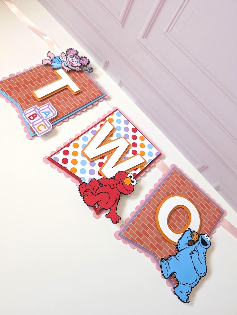 May include: A decorative banner spelling the word TWO, featuring three square panels with scalloped edges. The panels display the letters T, W, and O, decorated with Sesame Street characters Abby Cadabby, Elmo, and Cookie Monster on brick and polka dot backgrounds.