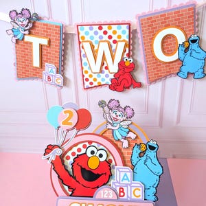 May include: A decorative party banner spelling TWO with Sesame Street characters and a matching cake topper featuring Elmo, Cookie Monster, and Abby Cadabby. The topper includes the name SIMONE, colorful balloons, and alphabet blocks in a layered design.