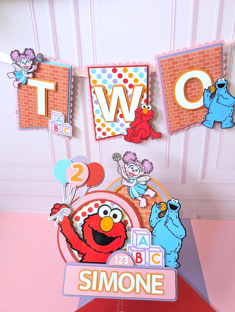 May include: A decorative party banner spelling TWO with Sesame Street characters and a matching cake topper featuring Elmo, Cookie Monster, and Abby Cadabby. The topper includes the name SIMONE, colorful balloons, and alphabet blocks in a layered design.