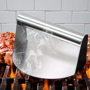 May include: A stainless steel grill cover is positioned over a piece of grilled meat on a barbecue grill. The cover is curved and reflects the light. Smoke rises from the meat, and flames are visible below the grill.