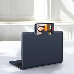 May include: A navy blue laptop with a smartphone attached to the top, displaying a video call. The phone is held in place by a small, matching blue holder. The laptop is open, and the screen is visible.