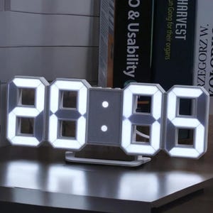 May include: A white digital LED clock displaying the time 28:06. The clock has a modern design with large, illuminated numbers and a small stand. The clock is on a dark surface, with books in the background.
