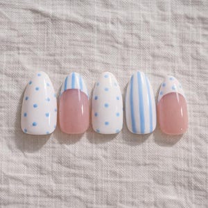 Op de afbeelding: Vijf kunstnagels met een lichtroze basis en verschillende ontwerpen. Twee nagels hebben witte bases met kleine blauwe stippen. Twee nagels hebben een lichtroze basis met een witte punt en blauwe strepen. Eén nagel heeft blauwe strepen.