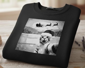 May include: Black sweatshirt featuring a grayscale graphic of a cat wearing a Santa hat, taking a selfie with a snowy winter landscape background. The image includes Santa's sleigh and reindeer.