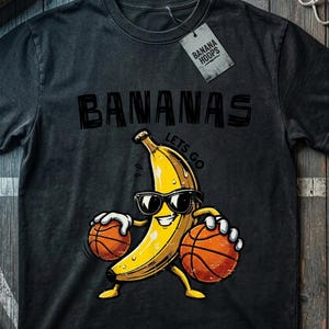 May include: Dark gray t-shirt with a graphic of a yellow banana wearing sunglasses and holding two basketballs. The word "BANANAS" is printed above the banana, and the tag reads "BANANA HOOPS".