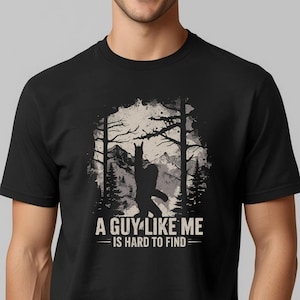 May include: Black t-shirt featuring a Bigfoot silhouette in a forest scene with mountains in the background. The text "A GUY LIKE ME IS HARD TO FIND" is printed below the graphic in white.