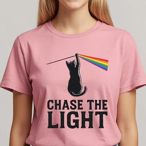 May include: Pink t-shirt featuring a black cat reaching for a rainbow prism. The text "CHASE THE LIGHT" is printed below the cat in a bold, black font. The shirt is a solid pink color.