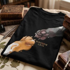 May include: Black t-shirt featuring a graphic of two cat paws touching, with claws extended. The text "WARRIOR CATS" is printed in gold. The shirt is displayed on a surface with books and a quill.