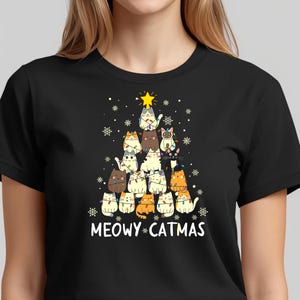 May include: Black t-shirt featuring a Christmas tree design made of various cartoon cats. The cats are decorated with string lights and topped with a yellow star. The text "MEOWY CATMAS" is printed below the tree.