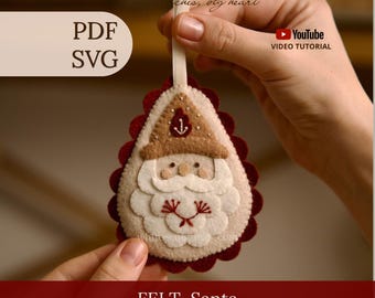 Può includere: Un ornamento in feltro a forma di goccia raffigurante Babbo Natale, con un cappello beige decorato da un'ancora, una barba bianca con ricami rossi e un supporto smerlato rosso scuro, dotato di un nastrino color crema per appenderlo.