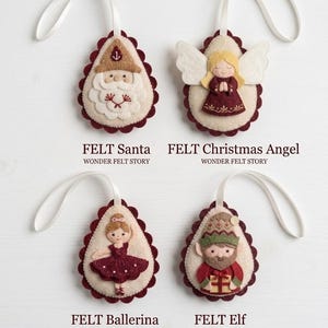 Puede incluir: Cuatro adornos de fieltro en forma de lágrima con cintas blancas. Los diseños incluyen un Santa, un ángel de Navidad, una bailarina y un elfo. Cada uno tiene un borde festoneado rojo oscuro y detalles bordados. El texto dice FELT Santa, FELT Christmas Angel, FELT Ballerina, FELT Elf y WONDER FELT STORY.