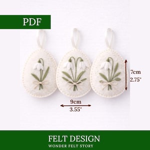 May include: Three off-white felt egg ornaments, each with a snowdrop design and a ribbon for hanging. The ornaments are 9cm wide and 7cm tall. The text "PDF" and "FELT DESIGN WONDER FELT STORY" are also visible.