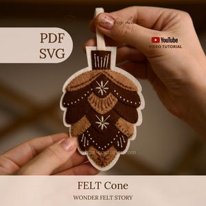 May include: A felt pinecone ornament featuring layered brown and tan fabric scales with white embroidered star and line details. The ornament includes a white felt hanging loop at the top. Text on the image includes PDF, SVG, YouTube Video Tutorial, FELT Cone, and WONDER FELT STORY.