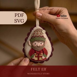 Könnte beinhalten: Ein tropfenförmiger Filzanhänger mit einem Elfenmotiv, der einen Bart und eine grün-beige Mütze trägt und ein goldenes Geschenk mit roter Schleife hält. Der Anhänger hat einen dunkelroten, gewellten Rand, eine cremefarbene Mitte und eine Schlaufe.