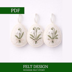 Op de afbeelding: Drie eivormige ornamenten van gebroken wit vilt met sneeuwklokjesontwerpen. Elk ei heeft een wit lint om op te hangen. De sneeuwklokjes zijn groen en wit, met een beige strik. De tekst "FELT DESIGN" en "WONDER FELT STORY" zijn zichtbaar.