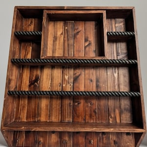 Op de afbeelding: Een rustiek houten wandrek met een donkere vlek. Het heeft een centraal verzonken gebied, twee planken en twee accenten in touwstijl. Het rek heeft een vierkante vorm en is geschikt voor het uitstallen van items.
