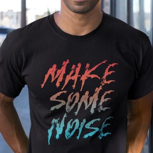 Könnte beinhalten: Schwarzes T-Shirt mit dem Schriftzug "MAKE SOME NOISE" in Rot-, Grau- und Blautönen. Die Person im Bild trägt eine schwarze Kappe mit grünem Schirm und farbigem Streifen. Die Person lächelt.