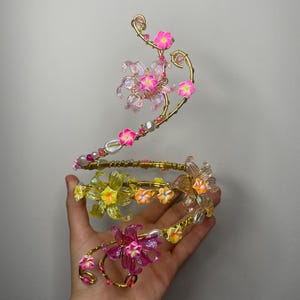 Op de afbeelding: Een bloemenarmband met een gouden draadbasis, versierd met roze, gele en oranje bloemen. De armband heeft transparante en roze kralen en witte parels. Het ontwerp is ingewikkeld en delicaat, geschikt voor speciale gelegenheden.