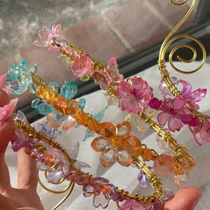 May include: Close-up of several decorative hair accessories. Each accessory features a gold wire base adorned with colorful, translucent flowers and beads in shades of pink, orange, blue, and purple. The design includes a swirl detail.