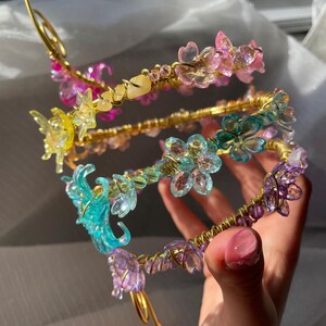 May include: A collection of floral headbands in various colors, including pink, yellow, blue, and purple. The headbands feature delicate, translucent flower and butterfly accents, and are crafted with gold-colored wire. The design is intricate and whimsical.