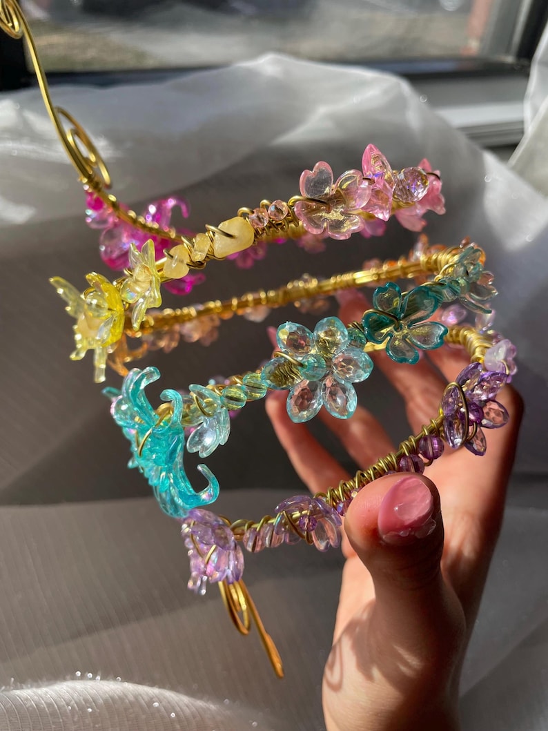 May include: A collection of floral headbands in various colors, including pink, yellow, blue, and purple. The headbands feature delicate, translucent flower and butterfly accents, and are crafted with gold-colored wire. The design is intricate and whimsical.