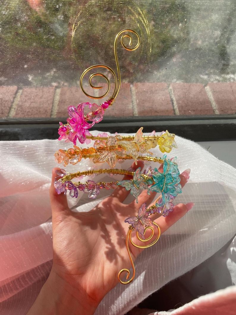May include: A decorative gold-colored wire sculpture with four tiers of colorful, translucent butterfly accents. The butterflies are pink, yellow, purple, and teal. The sculpture has a swirling design.