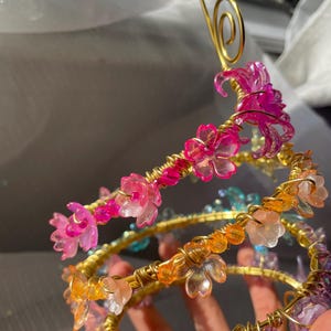 May include: A handcrafted, spiral-shaped headpiece with a gold wire frame. The headpiece is adorned with translucent pink, orange, and purple flower-shaped beads. A gold wire swirl tops the design. The headpiece is a unique accessory.