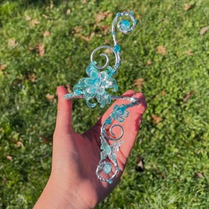 May include: A silver-toned, floral-themed ear cuff and matching bracelet set. The ear cuff features a large, translucent blue flower and a spiraling design with blue accents. The bracelet has smaller flowers and a similar design. The set is held against a green grassy background.
