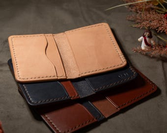 May include: A stack of three leather card holders in tan, navy blue, and brown. Each wallet features visible stitching along the edges and multiple interior slots for cards. The top tan wallet has a curved cutout on the left side.