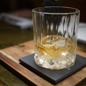 May include: A clear glass tumbler with a faceted, diamond-cut pattern on the lower half, containing a golden amber liquid and a single large ice cube. The glass sits centred on a square, dark grey slate coaster.