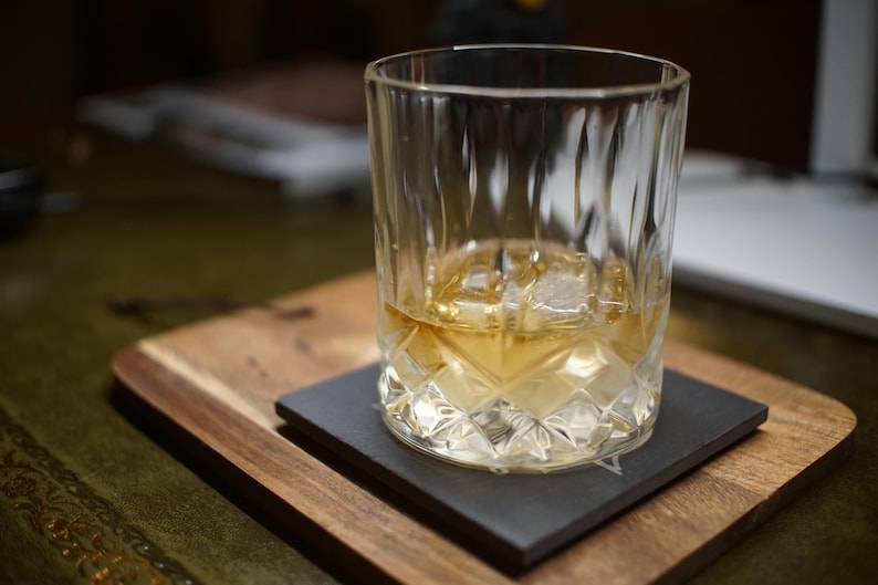 May include: A clear glass tumbler with a faceted, diamond-cut pattern on the lower half, containing a golden amber liquid and a single large ice cube. The glass sits centred on a square, dark grey slate coaster.