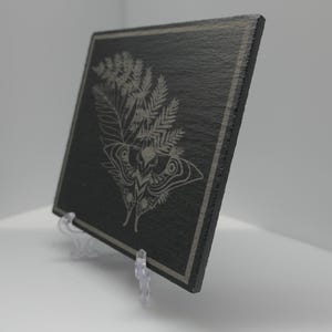 May include: A rectangular black slate plaque featuring a light grey laser-engraved design of a moth positioned beneath a large fern frond. The plaque has a thin border line etched around the perimeter and is displayed on a clear plastic stand.