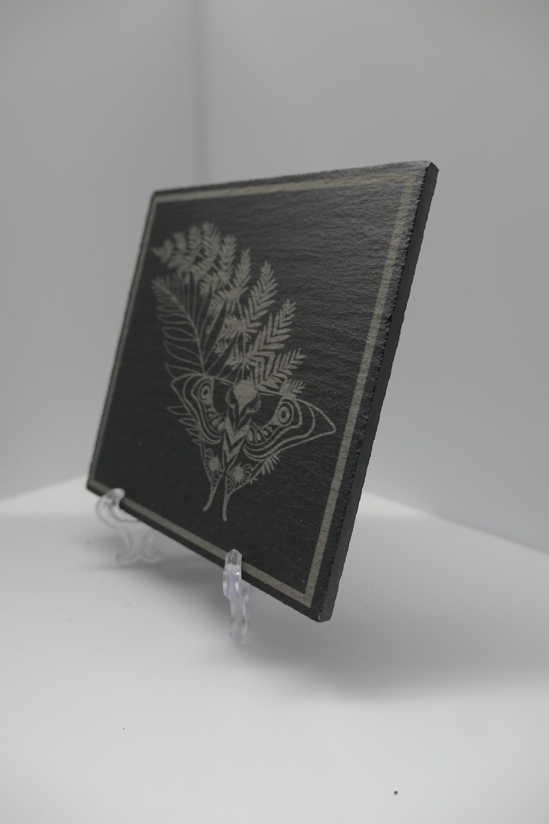 May include: A rectangular black slate plaque featuring a light grey laser-engraved design of a moth positioned beneath a large fern frond. The plaque has a thin border line etched around the perimeter and is displayed on a clear plastic stand.