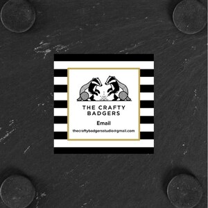 May include: A square business card featuring a black and white horizontal striped border. The centre white panel displays a gold frame, two badger illustrations, and the text: THE CRAFTY BADGERS, Email, thecraftybadgersstudio@gmail.com.